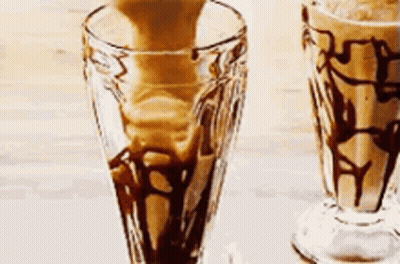 A Close Up Of Two Milkshakes In Glasses With Chocolate Sauce On A Table . GIF