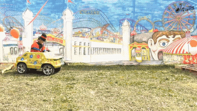A Clown Is Driving A Toy Car In Front Of A Cyclone Ride GIF
