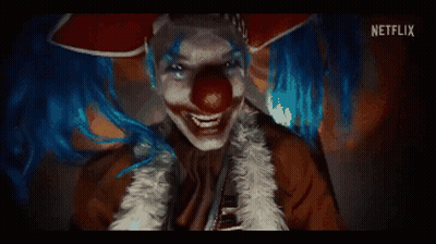 A Clown With Blue Hair And A Red Nose Is Smiling And Looking At The Camera . GIF