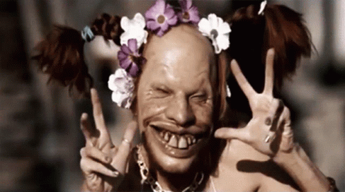 A Clown With Pigtails And Flowers On His Head Giving A Peace Sign GIF