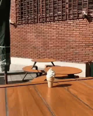 A Cone Of Ice Cream Is Sitting On A Wooden Table In Front Of A Brick Wall . GIF