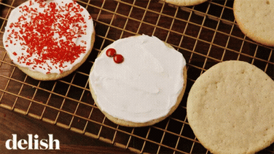 A Cookie With White Frosting And Red Sprinkles Sits On A Wire Rack With The Word Delish Below It GIF