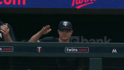 A Couple Of Baseball Players Sitting In A Dugout With A Twins Logo On The Wall GIF