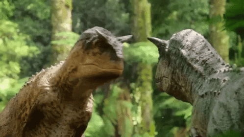 A Couple Of Dinosaurs Are Standing Next To Each Other In The Woods . GIF