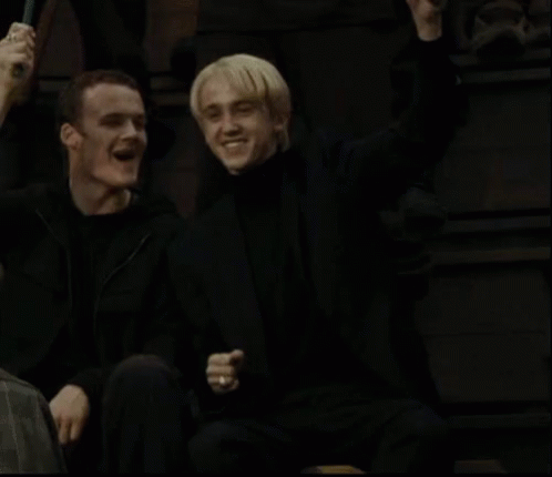 A Couple Of Men Are Sitting Next To Each Other In A Dark Room And Smiling . GIF