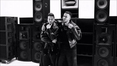 A Couple Of Men Are Standing Next To Each Other In Front Of A Stack Of Speakers . GIF