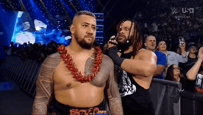 A Couple Of Men Standing Next To Each Other In A Wrestling Ring . One Of The Men Is Wearing A Lei . GIF