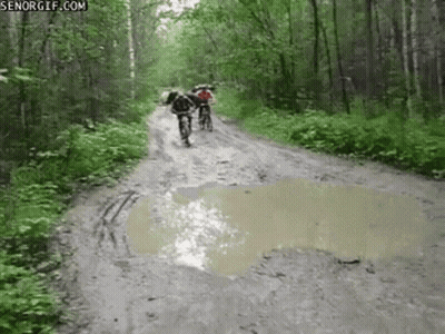 A Couple Of People Riding Bikes Down A Muddy Road . GIF