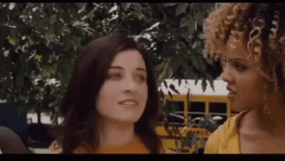 A Couple Of Women Are Standing Next To Each Other In Front Of A School Bus . GIF