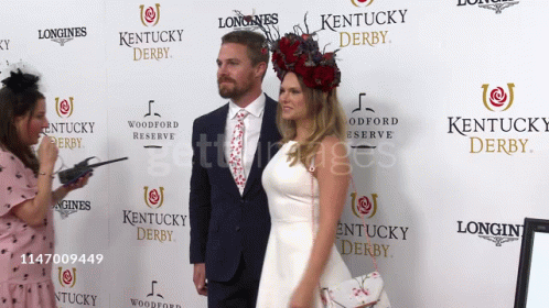 A Couple Standing In Front Of A Longines Kentucky Derby Sign GIF
