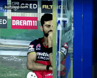 A Cricket Player Is Sitting In Front Of A Glass Door With His Hand On His Chin . GIF