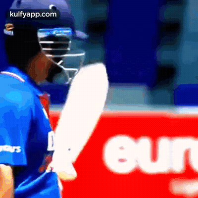 A Cricket Player Is Wearing A Helmet And Holding A Bat In Front Of A Red Sign . GIF