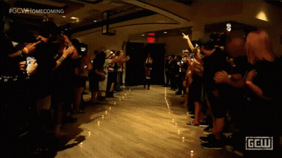 A Crowd Of People Are Gathered In A Dark Room With A Sign That Says Gcw Homecoming GIF