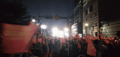 A Crowd Of People Holding Up Flags In Front Of A Building That Says ' Sydney Bank ' On It GIF