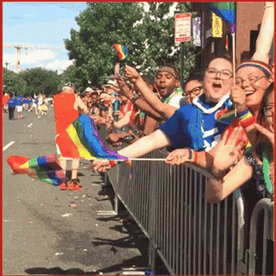 A Crowd Of People Watching A Parade Holding Rainbow Colored Flags GIF