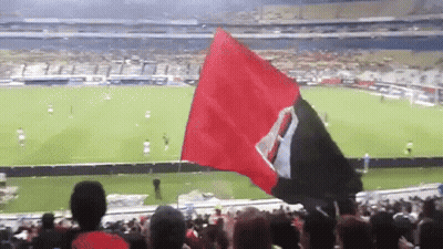 A Crowd Of People Watching A Soccer Game With A Red And Black Flag Flying In The Foreground GIF