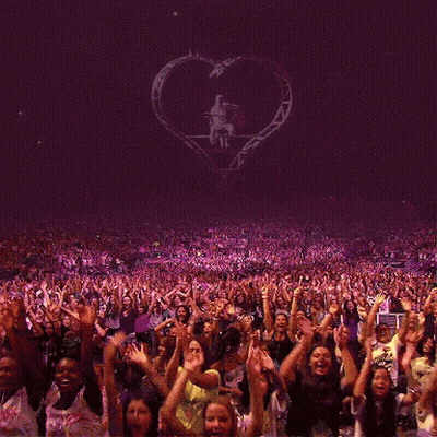A Crowd Of People With Their Hands In The Air At A Concert With A Heart In The Background GIF