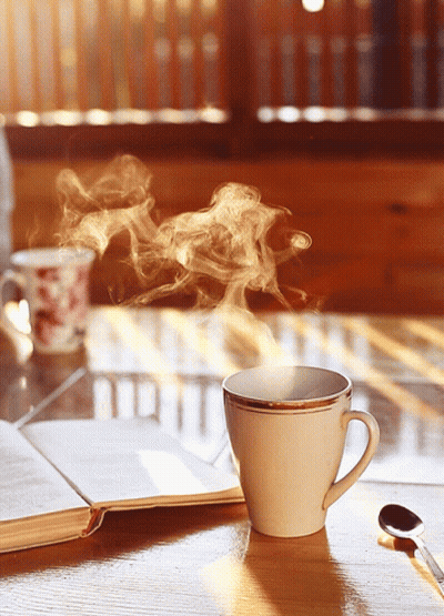 A Cup Of Coffee With Steam Coming Out Of It Sits On A Table Next To An Open Book GIF