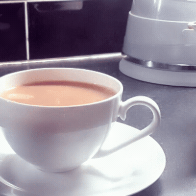A Cup Of Tea Sits On A Saucer Next To A White Kettle GIF