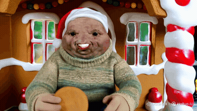 A Doll Wearing A Santa Hat And A Sweater Is Sitting In Front Of A Gingerbread House GIF