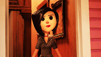 A Doll With Black Hair And Red Lips Stands In Front Of A Pink House GIF