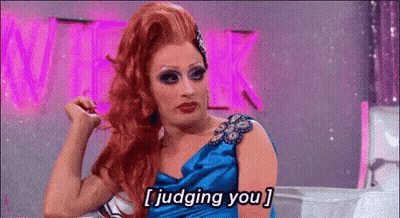 A Drag Queen In A Blue Dress Is Sitting In Front Of A Pink Sign And Says `` Judging You '' . GIF