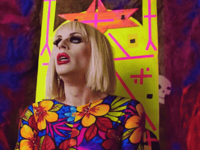 A Drag Queen In A Colorful Dress Stands In Front Of A Neon Sign With A Cross On It GIF