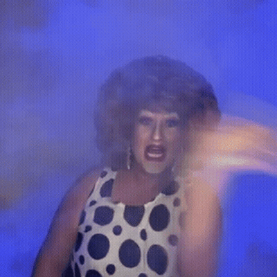A Drag Queen In A Polka Dot Dress Is Standing In Front Of A Blue Background And Making A Funny Face . GIF