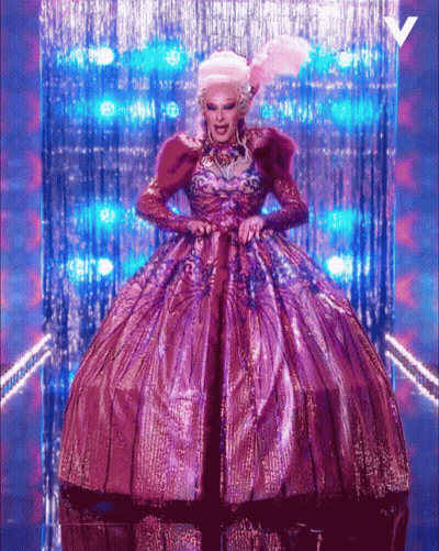 A Drag Queen In A Purple And Gold Dress With A V On The Bottom GIF