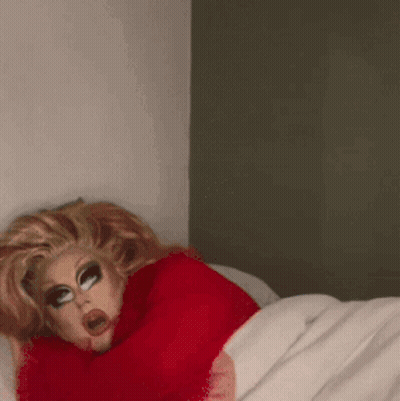 A Drag Queen In A Red Sweater Is Laying In Bed GIF