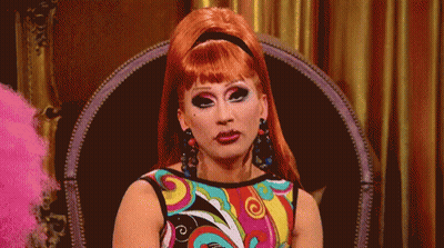 A Drag Queen Is Sitting In A Chair Wearing A Colorful Dress GIF