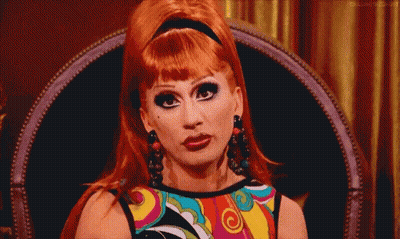 A Drag Queen Is Sitting In A Chair Wearing A Colorful Shirt And Earrings . GIF