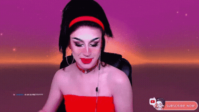 A Drag Queen Is Sitting In Front Of A Purple Background And Holding A Cotton Candy Bar . GIF