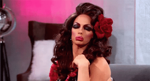 A Drag Queen Is Sitting On A Couch With A Red Flower In Her Hair . GIF