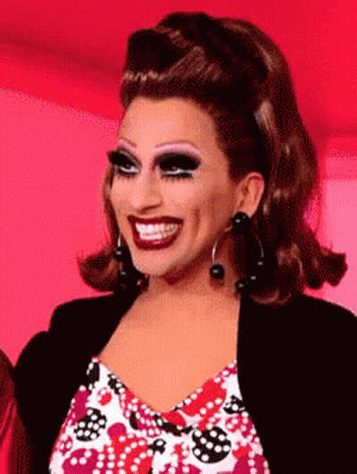 A Drag Queen Is Smiling And Wearing A Black Jacket And Earrings GIF