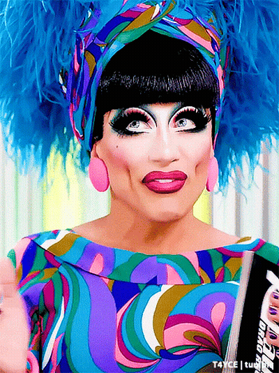 A Drag Queen Is Wearing A Colorful Dress And Feathered Headpiece GIF