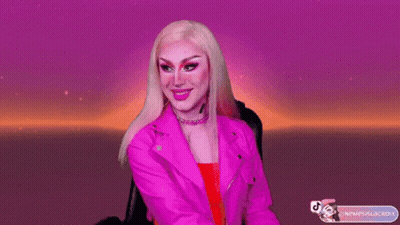 A Drag Queen Is Wearing A Pink Jacket And Earrings GIF