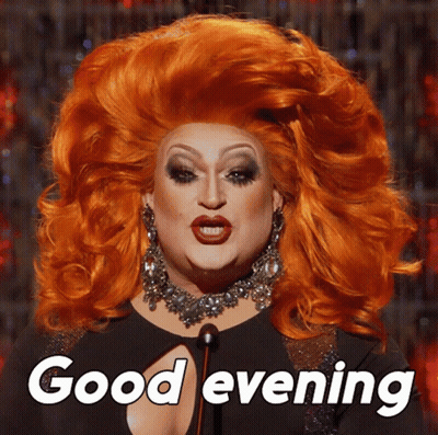A Drag Queen Says " Good Evening " While Wearing A Large Red Wig GIF