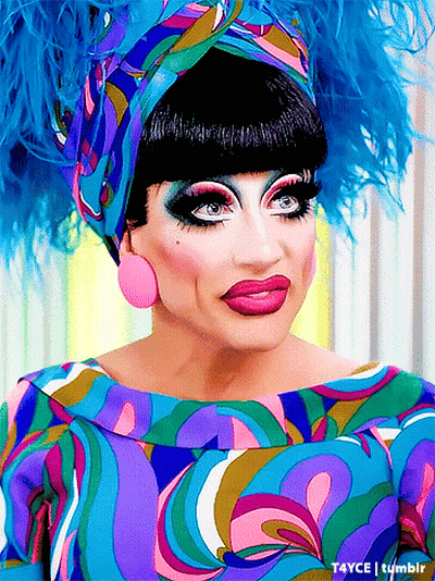 A Drag Queen Wearing A Colorful Dress And Feathered Headpiece GIF