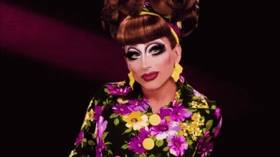 A Drag Queen Wearing A Colorful Floral Shirt And Yellow Earrings GIF
