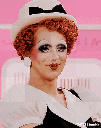 A Drag Queen Wearing A White Hat And Earrings GIF