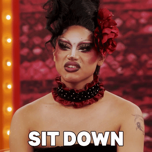 A Drag Queen With A Red Flower In Her Hair Is Sitting Down GIF