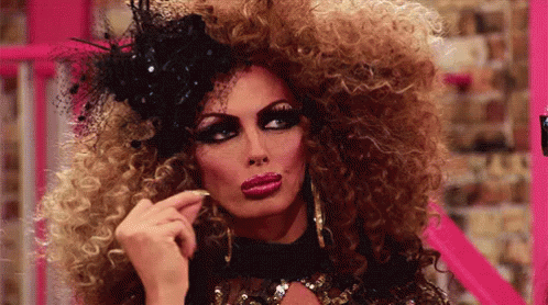 A Drag Queen With A Very Large Curly Hair And A Black Hat GIF