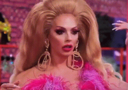 A Drag Queen With A Very Large Wig And Earrings Is Wearing A Pink And Purple Outfit . GIF