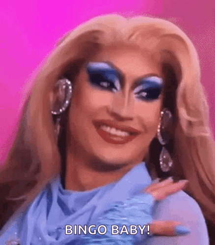 A Drag Queen With Blonde Hair And Blue Makeup Is Smiling And Making A Bingo Baby Gesture . GIF