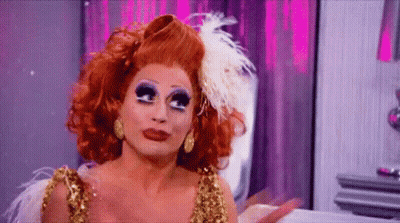 A Drag Queen With Red Hair And Feathers On Her Head Is Wearing A Gold Dress And Earrings . GIF