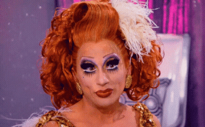 A Drag Queen With Red Hair And Feathers On Her Head Looks At The Camera GIF