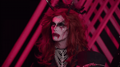 A Drag Queen With Red Hair And Horns Is Wearing A Pearl Necklace GIF