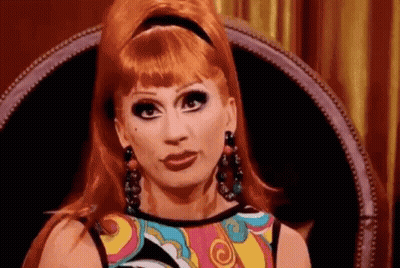 A Drag Queen With Red Hair Is Sitting In A Chair And Making A Funny Face . GIF