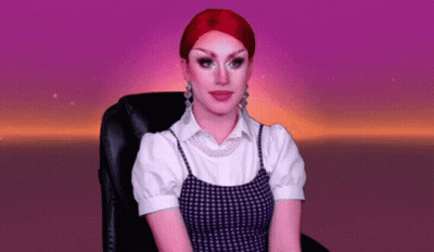 A Drag Queen With Red Hair Is Sitting In A Chair With A Purple Background . GIF
